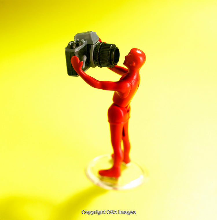 Red Figurine Model Holding Camera on Yellow Background in Artistic Setup