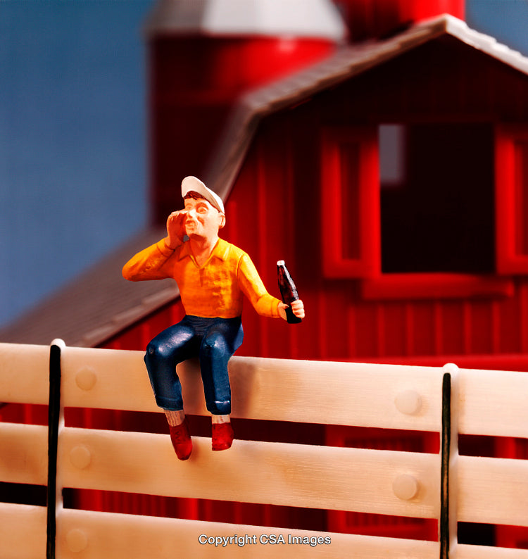 Plastic Figure Sitting on Fence by Barn Holding a Bottle